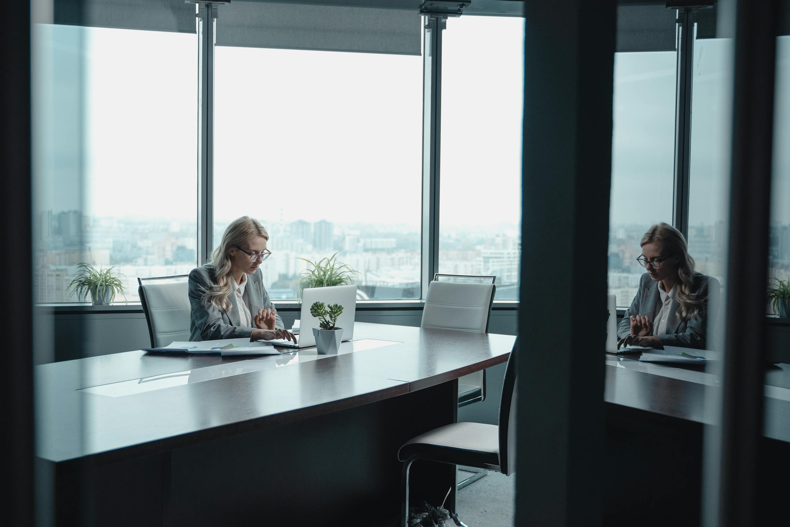 A businesswoman in a blazer works at a desk in a modern office with city views.