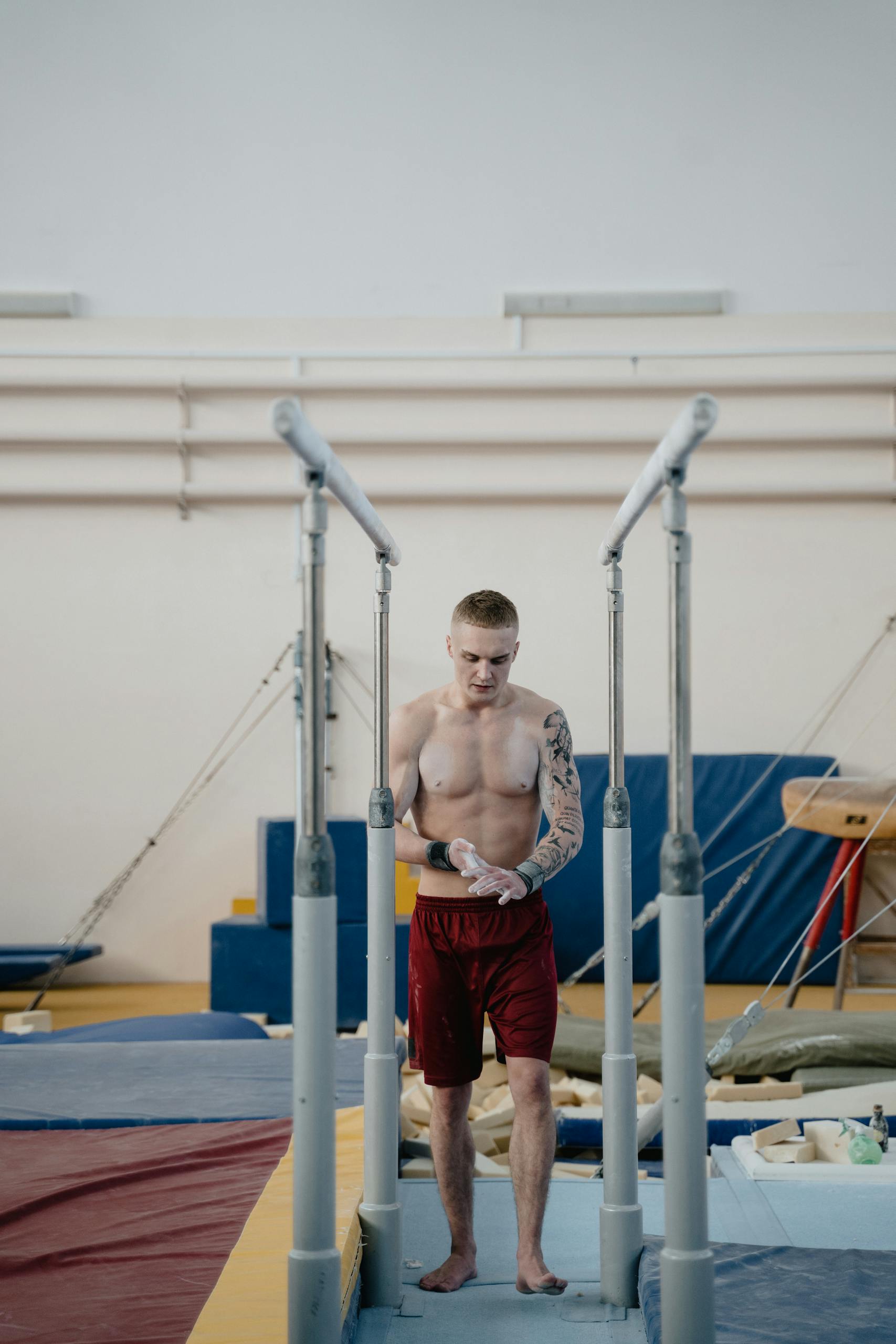 A shirtless man practicing exercises on parallel bars in an indoor gym setting.