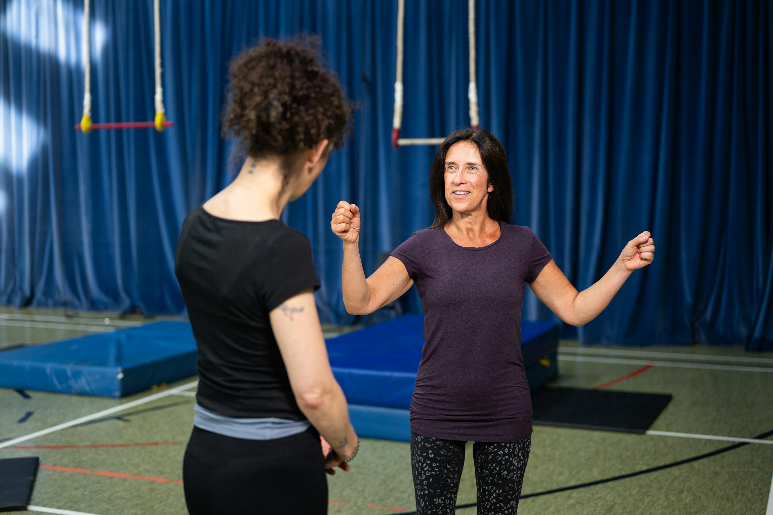 Two women engaged in a trapeze class inside a gym with mats and ropes.